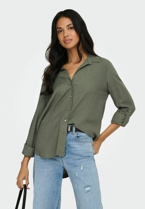 Femme aux longs cheveux noirs portant une chemise olive à manches retroussées et un jean bleu clair déchiré, tenant un sac bandoulière noir.