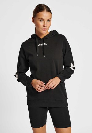 Jonge vrouw draagt een zwarte Hummel hoodie met wit logo en zwarte fietsbroek, staat met gevouwen handen voor zich.