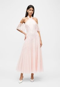 Γυναίκα που φοράει ένα ανοιχτό ροζ φόρεμα με λαιμόκοψη halter, διαφανείς φουσκωτούς ώμους εκτός ώμων και υφάσματα με ανάγλυφα λουλούδια, συνδυασμένο με λευκά μυτερά τακούνια.