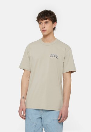 Camiseta de algodón color beis con cuello redondo, mangas cortas y un pequeño logo negro de "Dickies" en el lado izquierdo del pecho. Textura suave, corte relajado.
