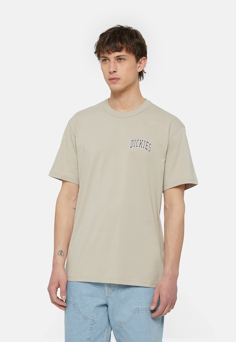 Bežīga kokvilnas T-kreklu ar apaļu kaklu, īsām piedurknēm un mazu melnu "Dickies" logotipu kreisajā krūtī. Gluda tekstūra, brīvs piegriezums.