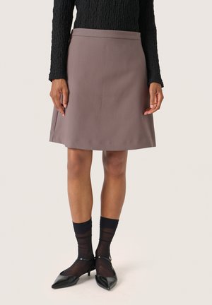 Une jupe A-line de longueur genoux en tissu brun muted avec une texture lisse. Portée avec des chaussettes noires transparentes et des escarpins noirs pointus.