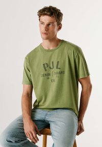 Olivovozelené bavlnené tričko s okrúhlym výstrihom a krátkymi rukávmi, s potlačeným logom v tmavo šedej farbe. Nosené s svetlomodrými džínami.