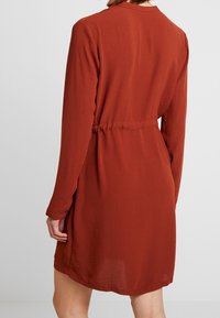 Ruskottinen pitkähihainen mekko, jossa on istuva vyötärö, suora leikkaus ja hienovarainen tekstuuri. Ominaisuuksiin kuuluvat korkea kaula-aukko ja vähäinen yksityiskohtaisuus.