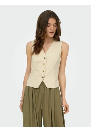 Femme aux cheveux bruns ondulés portant un gilet beige sans manches boutonné et un pantalon ample vert olive, regardant vers le bas avec les bras détendus.