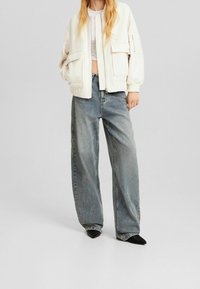 Biała kurtka bomberka na zamek błyskawiczny z dużymi kieszeniami, zestawiona z luźnymi, wyblakłymi niebieskimi spodniami o szerokich nogawkach i czarnymi butami na szpic.