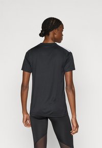 Μαύρο αθλητικό μπλουζάκι με κοντά μανίκια, λείο ύφασμα, ελαφριά καμπύλη στο κάτω μέρος και λευκή λεπτομέρεια κατά μήκος των ώμων.