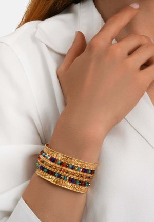 Bracelet manchette en or avec des motifs complexes et des perles multicolores porté au poignet contre la manche d'une chemise blanche.