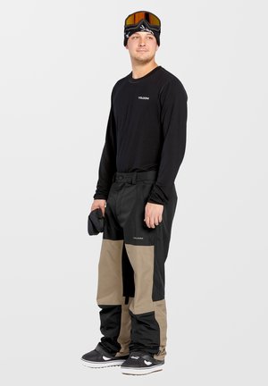 Camisa negra de manga larga con logo, pantalones impermeables en tonos marrón y negro, zapatillas negras, usando gafas y un gorro de lana, manos sosteniendo un guante.
