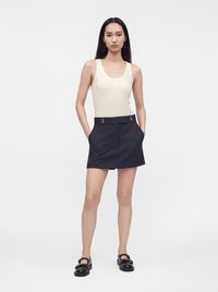 Femme aux longs cheveux foncés portant un top blanc sans manches, une jupe noire taille haute avec des boucles, et des chaussures noires, debout les mains dans les poches.