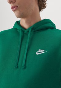Vihreä puuvillainen huppari, jossa on teksturoidun näköinen pinta. Siinä on etutasku, kiristysnauhalla varustettu huppu ja valkoinen Nike-logo rinnassa.