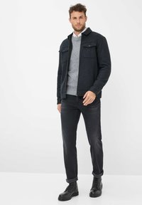Giacca zip grigia scura con due tasche sul petto, indossata sopra un maglione grigio chiaro. I jeans in denim neri e gli stivali neri con lacci completano l'outfit.
