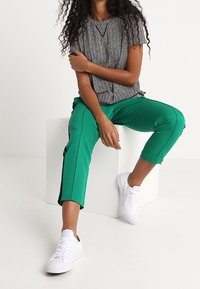 Top de patrón zigzag en blanco y negro combinado con pantalones verdes ajustados con rayas laterales. Usado con zapatillas blancas; modelo sentado.