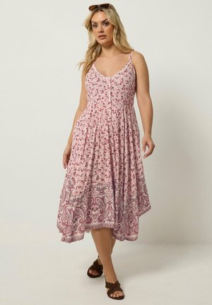 Kvinde med blondt hår iført en ærmeløs, lyserød og hvid blomster-midi kjole med asymmetrisk kant og brune sandaler.