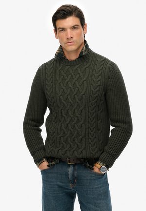 Dunkelgrüner Kabelstrickpullover mit hohem Kragen, strukturiertem Design und gerippten Ärmeln, getragen über einem karierten Hemd, kombiniert mit blauen Jeans.