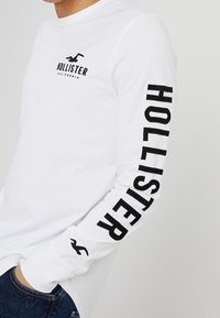 Vit långärmad t-shirt med svart text "HOLLISTER" längs den högra ärmen och en logotyp på bröstet; av bomull med rundad halsringning.