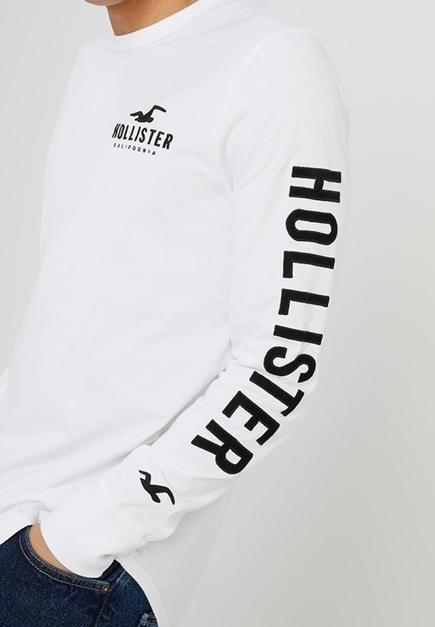 Vit långärmad t-shirt med svart text "HOLLISTER" längs den högra ärmen och en logotyp på bröstet; av bomull med rundad halsringning.