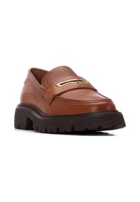 Mocasín de cuero marrón con punta redondeada, que presenta un acento metálico y una suela de goma negra gruesa con un diseño de banda de rodadura texturizada.