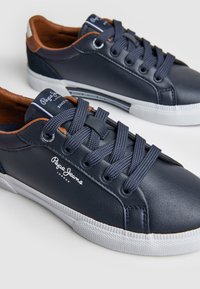 Tumsas zila Pepe Jeans ādas sporta apavi ar baltiem zoliem, biezām tumši zilām šņorēm un brūnu iekšpusi uz balta fona.