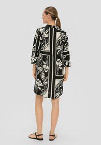 Robe noire et blanche à mi-longueur avec des manches trois-quarts, présentant un motif floral et un dos fendu. Portée avec de simples sandales noires.