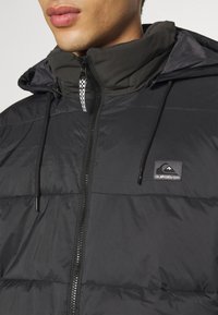 Black puffer jacket with a hood, featuring a zip closure, drawstring accents, and a logo patch. Smooth finish with a quilted texture.