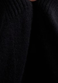 Musta neulottu kangas, jossa on joustinneuloksen rakenne, ja siinä on kodikas, paksu kutomus. Lähikuva, joka näyttää materiaalin pehmeyden ja syvyyden.