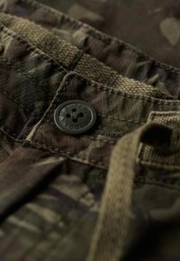 Détail de tissu camouflage avec un bouton sombre gravé "Superdry", couture piquée, ceinture texturée et cordon de serrage.