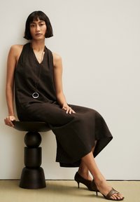 Femme aux cheveux noirs courts portant une robe sombre sans manches et un collier pendentif, assise sur un tabouret sculptural noir moderne, jambes croisées.
