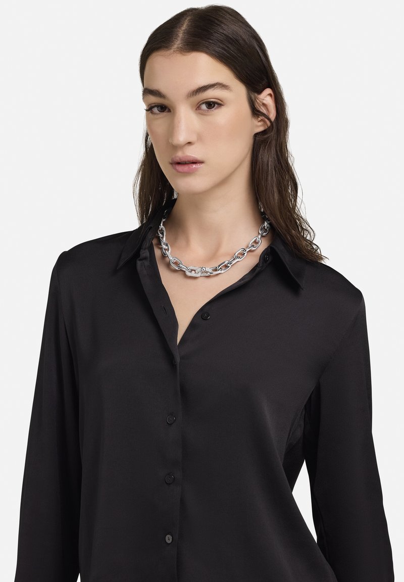 Camisa negra de manga larga en tejido liso con botones al frente y cuello con solapa, combinada con un collar de cadena plateada con eslabones.