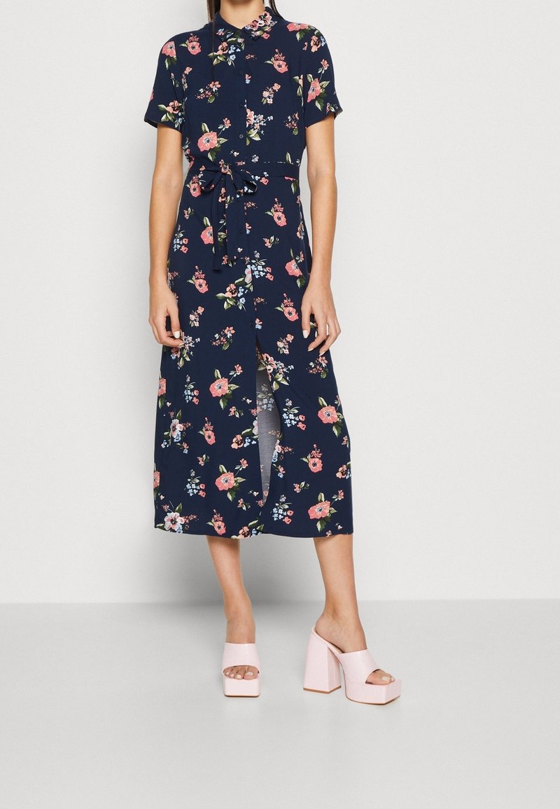 Robe florale marine à manches courtes et taille cintrée, avec des motifs floraux roses et verts, détail fente, associée à des talons blocs rose clair.