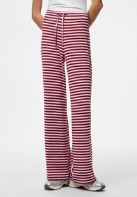 Person tr ägt weit geschnittene Hose mit horizontalen Streifen in Burgund und Rosa, kombiniert mit silbernen Turnschuhen, steht vor einfachem Hintergrund.