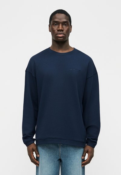 Νεαρός με σκούρο δέρμα, φοράει μπλε σκούρο πουκάμισο με μακριά μανίκια και ανοιχτόχρωμο τζιν, στέκεται μπροστά σε απλό λευκό φόντο.