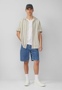 Kurzärmeliges, gestreiftes Hemd in Beige und Grün, kombiniert mit blauen Denim-Shorts. Darunter ein weißes T-Shirt, weiße Sneakers und eine graue Cap.