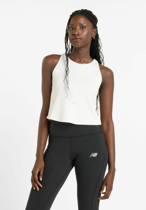 Femme aux cheveux tressés portant un haut blanc sans manches et des leggings noirs New Balance, debout devant un fond blanc uni.