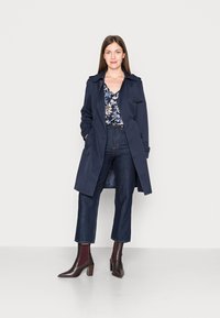 Manto de marinheiro com botões duplos, ombreiras e bolsos. Usado sobre uma blusa floral e combinado com jeans escuros e botins borgonha.