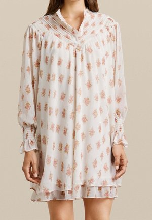 Woman wearing a white long-sleeve floral dress with pink and beige flower patterns and ruffled cuffs and hem.