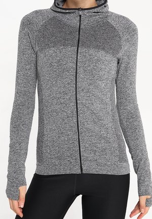 Grijze atletische hoodie met volledige ritssluiting en textuurpanelen op de schouders en zijkanten, gedragen met zwarte atletische leggings, handen ontspannen langs het lichaam.