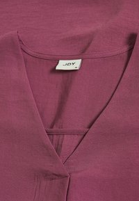 Bluzka z dekoltem w serek w głębokim kolorze mauve, wykonana z lekkiego materiału. Posiada metkę z napisem "JDY" i rozmiarem "36" na dekolcie.
