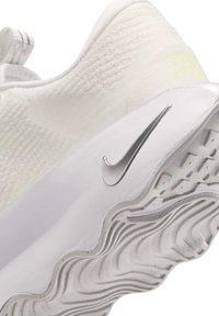 Gros plan d'une sneaker blanche Nike montrant un tissu texturé, le swoosh argenté sur le côté et une semelle à motifs ondulés.