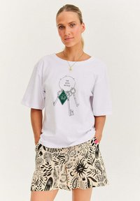 Kobieta nosząca białą koszulkę z grafiką breloczka i napisem "Klucze ujawniają", w połączeniu z beżowymi szortami w czarne abstrakcyjne wzory przyrody.