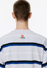 Harmaa raidallinen t-paita, jossa on sinisiä ja mustia raitoja, ja takana kaula-aukon lähellä pieni monivärinen PlayStation-logo.