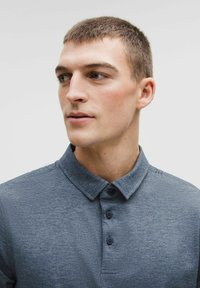 Młody mężczyzna z krótkimi brązowymi włosami, ubrany w niebieską koszulę polo z guzikami, patrzący w lewo na jednolity jasny tle.