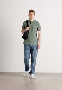 Ung mand går, iført en grøn polo-shirt, blå jeans, hvide sneakers og bærer en sort crossbody-taske på en ensfarvet baggrund.