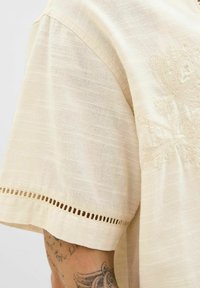 Top de manga corta beige hecho de una tela ligera, con delicados bordados florales y un patrón texturizado, con un detalle de encaje en el dobladillo.