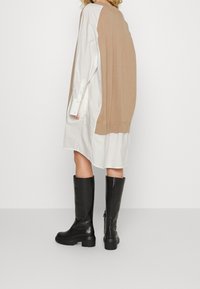 Pull en tricot beige surdimensionné porté par-dessus une chemise blanche, assorti de bottes en cuir noir montantes avec des semelles épaisses.