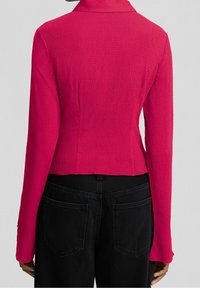 Blazer rosa de manga larga ajustado, tejido texturizado, cuello puntiagudo, diseño corto, combinado con jeans negros de cintura alta, con bolsillos visibles.