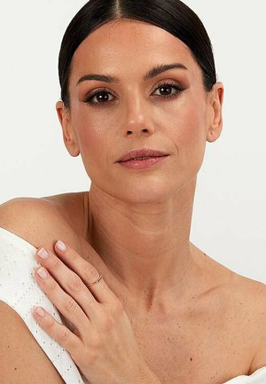 Close-up van het gezicht van een vrouw met lichte make-up, een gladde huid en donker haar dat opgestoken is. Ze draagt een witte top zonder schouderbandjes en een delicate ring.
