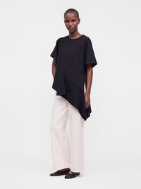 Femme portant un haut asymétrique noir à manches courtes, un pantalon blanc à jambes larges et des chaussures plates noires, debout devant un fond uni.