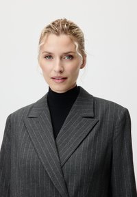 Blazer gris à rayures avec un design structuré, des revers larges et une texture douce, associé à un turtleneck noir en dessous.