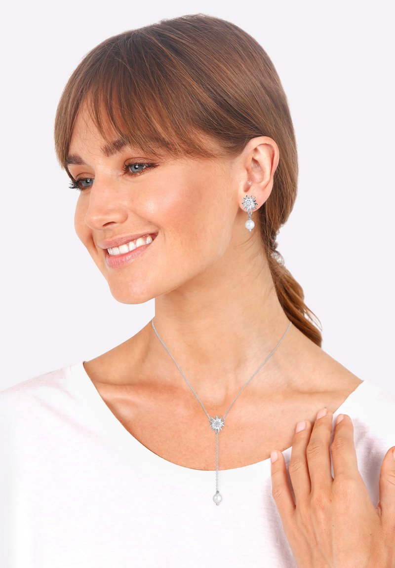 Silbernes Schmuckset mit blütenförmigen Ohrringen mit Perlen und einer langen Anhänger-Kette im ähnlichen Design. Getragen mit einem weißen Oberteil.